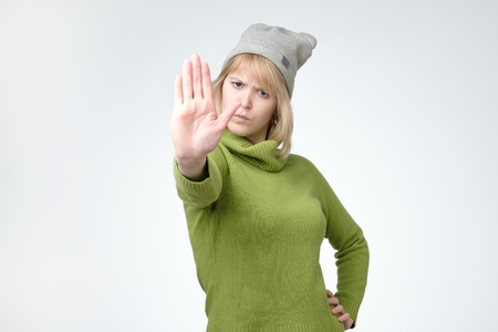 Young serious pretty girl in green sweater and hat showing stop sign. Prohibition symbolの写真素材