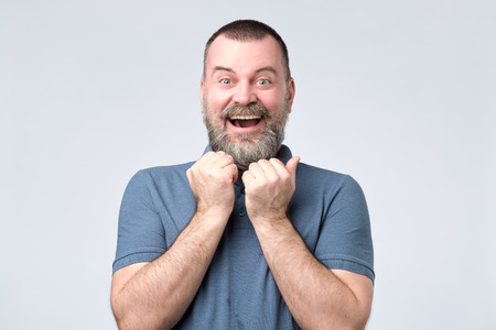 Excited bearded man standing with fists up on gray background.