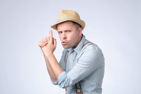 Young handsome man wearing hat and suspenders holding symbolic gun with hand gesture