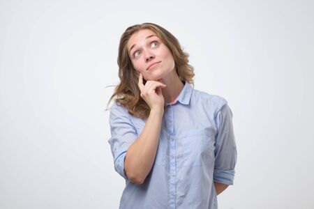 Pretty thoughtful caucasian young woman looks with doubt upwards, wears blue shirt. Pensive youngster being deep in thoughtsの写真素材