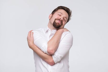 PLeased handsome bearded young man hugging himself with closed eyes and smile enjoying.の写真素材