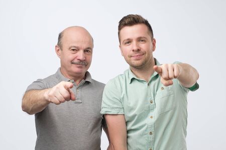 Two happy caucasian men friends or father and son hugging each other, pointing finger and looking at...