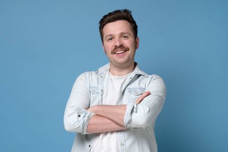 Smiling young man standing with arms crossed against blue background. Studio shot. Positive facial...