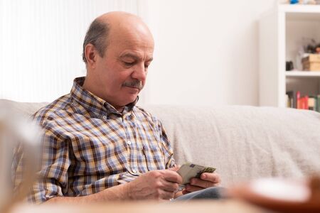 Senior bald man with mustache busy counting money and bills at home, sitting at home alone.