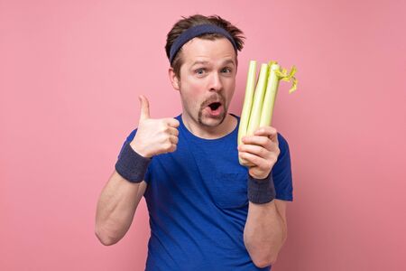 Young caucasian sportive trainer and nutritionist holding a green celery showing thumb up. Healthy diet. Studio shot on pink background.の写真素材