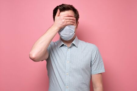 Caucasian young man wearing medical mask and covering eyes with hand trying to protect from flu or coronavirus. Studio shot on pink wall.の写真素材