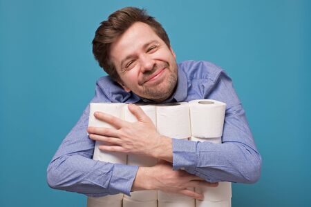 Excited happy caucasian man hugging a pile of toilet paper isolated on blue background. Studio shot