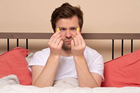 Caucasian young stressed man holding a yellow earplug trying to sleep having insomnia. Studio shot