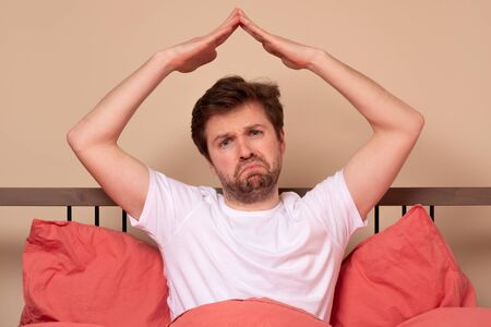 Caucasian young man in the bed showing roof of house gesture meaning coronavirus quarantine or staying at home.の写真素材