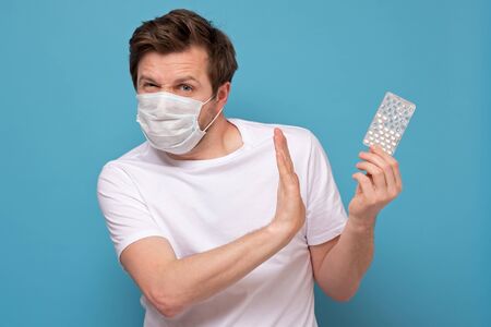 Caucasian man in medical mask with pills has a sore throat pointing on them and showing refusing gesture. Is it placebo concept.の写真素材