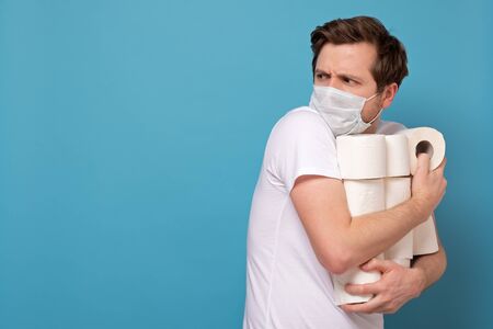Greed and panic because of coronavirus. Man in medical mask holding a lot of rolls of toilet paper hiding them from everybody. Studio photo on blue wallの写真素材