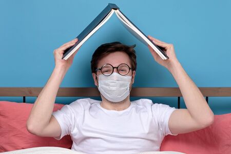 Caucasian young man in glasses and medical mask reading a novel in his bedroom being alone in...