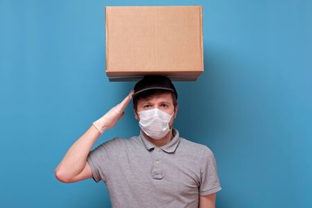 Young delivery man in medical mask holding and carrying a cardbox making salute ready for work on it isolated on blue backgroundの写真素材