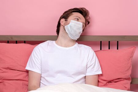 Young worried sick man in medical mask sitting on bed at home scary and desperate suffering...