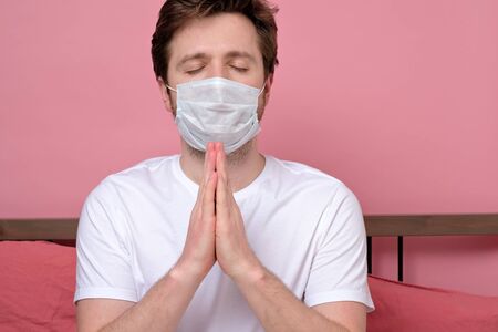 Man in medical mask holding hands together and praying while lying in bed aloneの写真素材