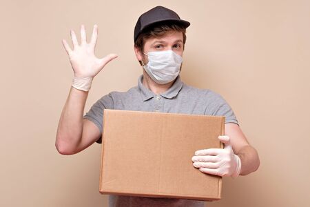 Young delivery man in medical mask showing five finger holding and carrying a cardbox isolated on...