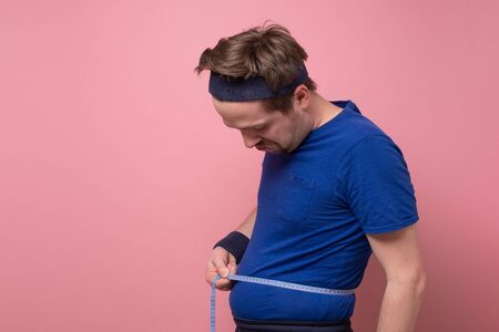 Funny caucasian young man measuring belly fat itself being sad with his diet results. Studio shot on...