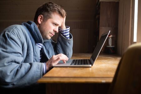 Handsome young man checks mail on a laptop sitting near window.