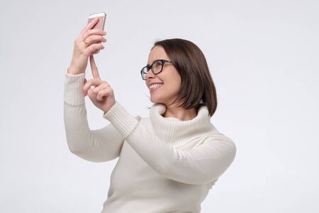 Mature smiling woman with glasses taking selfie or having online conversation on phone during quarantine. Studio shot on white wall.の写真素材