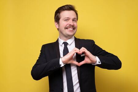 Happy young man in suit with mustache showing heart symbol. Studio shot on yellow wall.の写真素材