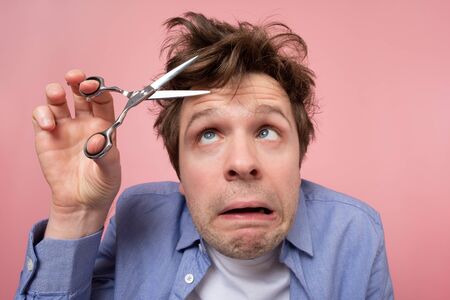 Caucasian shaggy man trying to make a hairstyle by himself with scissors being on quarantine. Studio...