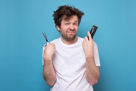Do it yourself. Caucasian shaggy man holding scissors and hair clipper is afraid to cut his hair during quarantine. Studio shot on blue wall.の写真素材