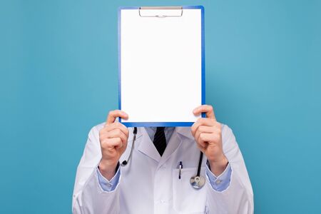 Young man as doctor in white coat and with stethoscope showing blank signboard with empty copyspace area for slogan or advertising text message. Studio shot on blue wallの写真素材