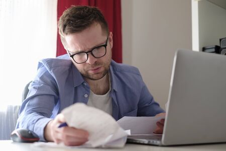 Young caucasian man working with laptop at home browsing bills and documents. Businessman going through paperwork at home office. Stay home and self isolation quarantine.の写真素材
