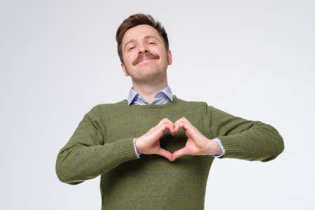 Young caucasian man with funny mustachedoing a heart gesture against a pink backgroundの写真素材