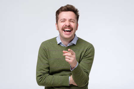 Young man with mustache pointing at camera and laughing, making fun towards you. Studio shotの写真素材