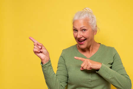 Senior woman pointing aside on copyspace. Studio shot on yellow wall.