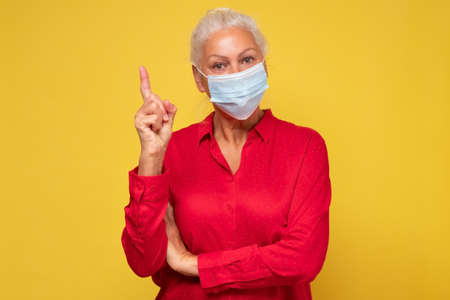 Senior woman wearing protective medical mask for protection from virus pointing index finger upの写真素材