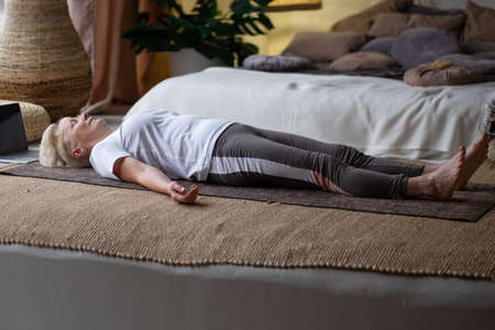 Senior woman working out at home, doing yoga exercise lying in Shavasana or Corpse or Dead Body Postureの写真素材