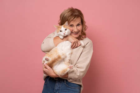 Mature woman lovingly hugging cat on pink wall.の写真素材
