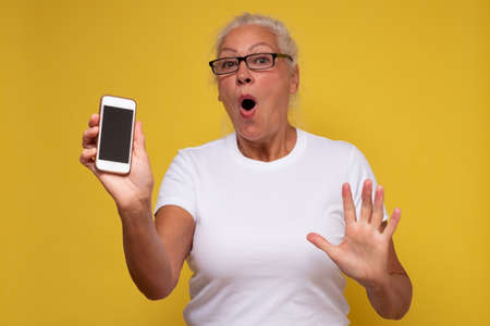 Woman sharing useful app. Mature caucasian mom wear glasses extend arm hold smartphone show phone screen display on yellow wall.の写真素材