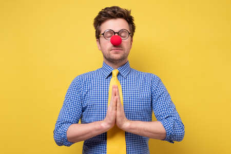 Funny young man with red clown nose pressings palms together, trying to calm down.の写真素材