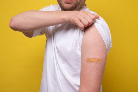 Caucasian man showing his vaccinated arm. He received a corona vaccine looking at camera. Studio shot on yellow wall.の写真素材