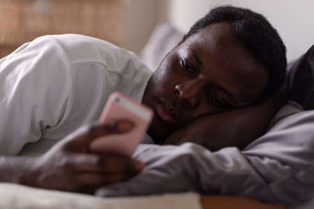 Sad african man lying in bed reading sms from his girlfriend