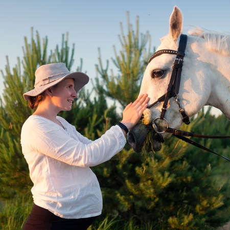 young pregnant woman feeding a white horse in the field