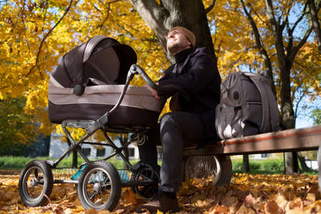 Happy young father with pram sitting on bench in nature at parkの写真素材