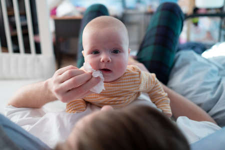 Young fatherwipes drool with white paper handkerchief of his newborn baby girl in bed.の写真素材