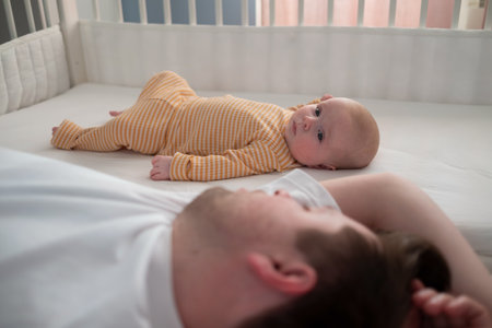 Tired father sleeping and his newborn son lying on bed looking at camera at home.の写真素材