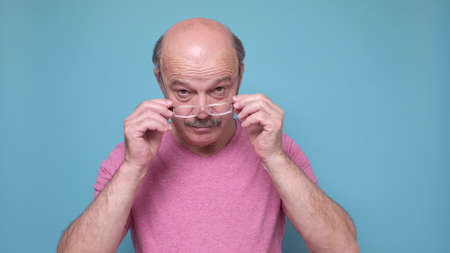 Senior hispanic man looking through glasses being shocked. Studio shot on blue wall.の写真素材
