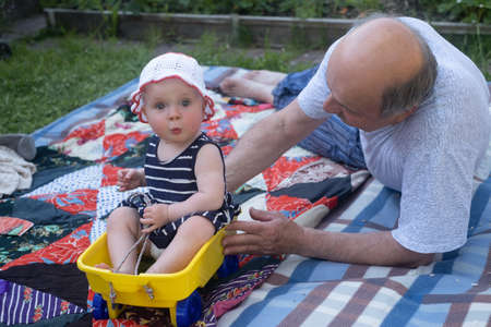 Grandfather playing with his grandchild on blanket outdoors pushing small cart.の写真素材