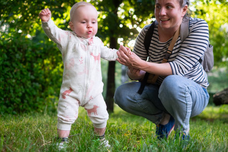 Mom learning baby daughter to walk, first steps in park at summer warm day.の写真素材
