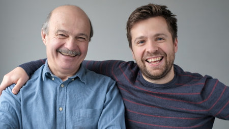 Happy mature senior father and grown son standing together, hugging, looking at camera