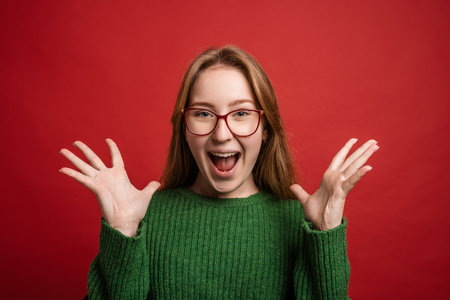 A young woman with long hair and glasses, wearing a green sweater, expresses excitement with her hands raised and a big smile against a red background.の写真素材
