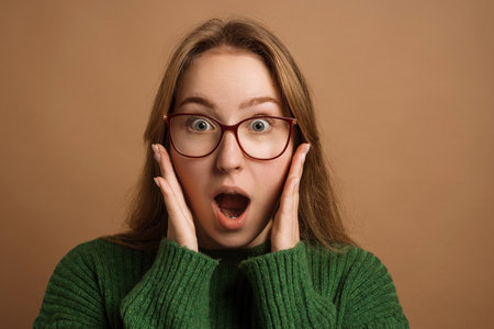 A surprised young woman with glasses, wearing a green sweater, holds her face in shock against a brown background.の写真素材