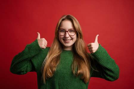 portrait of a happy girl in glasses holding thumbs up on red backgroundの写真素材