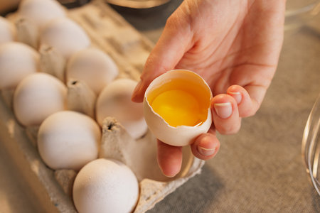 A person holding a cracked eggshell with a bright yellow yolk, surrounded by a tray of whole eggs....